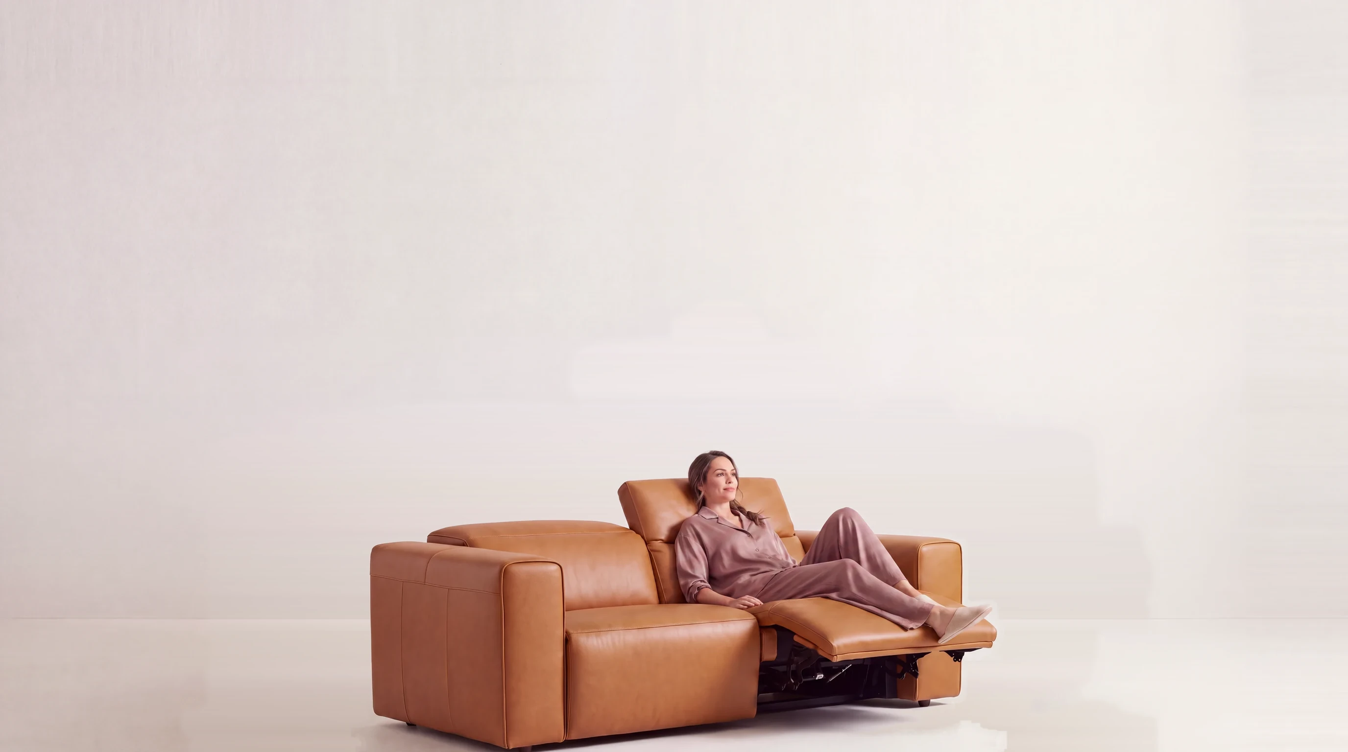 A woman reclining on a tan leather Sofa Studio electric recliner sofa, one seat reclined with the footrest extended and the headrest raised